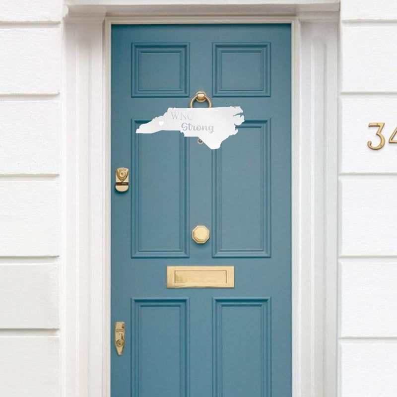 Blue door with gold handle and mail slot, featuring a decorative steel sign shaped like the state of North Carolina with "WNC Strong" Purchase online today at Harvest Array.